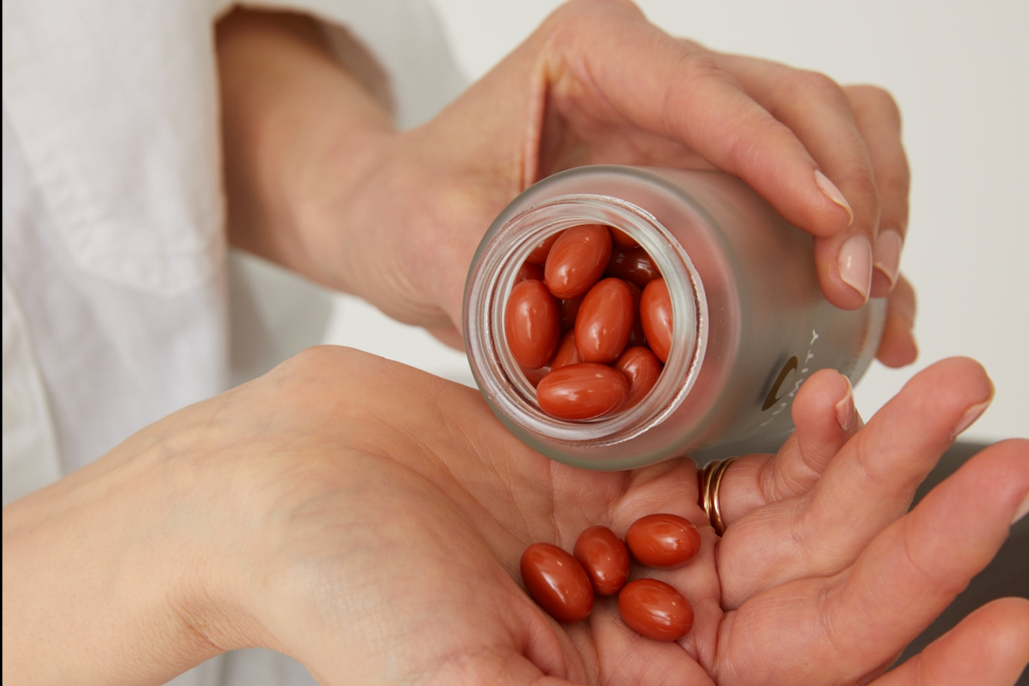 Close-up of hands holding an open bottle pouring reddish wellness supplements into the palm against a neutral background