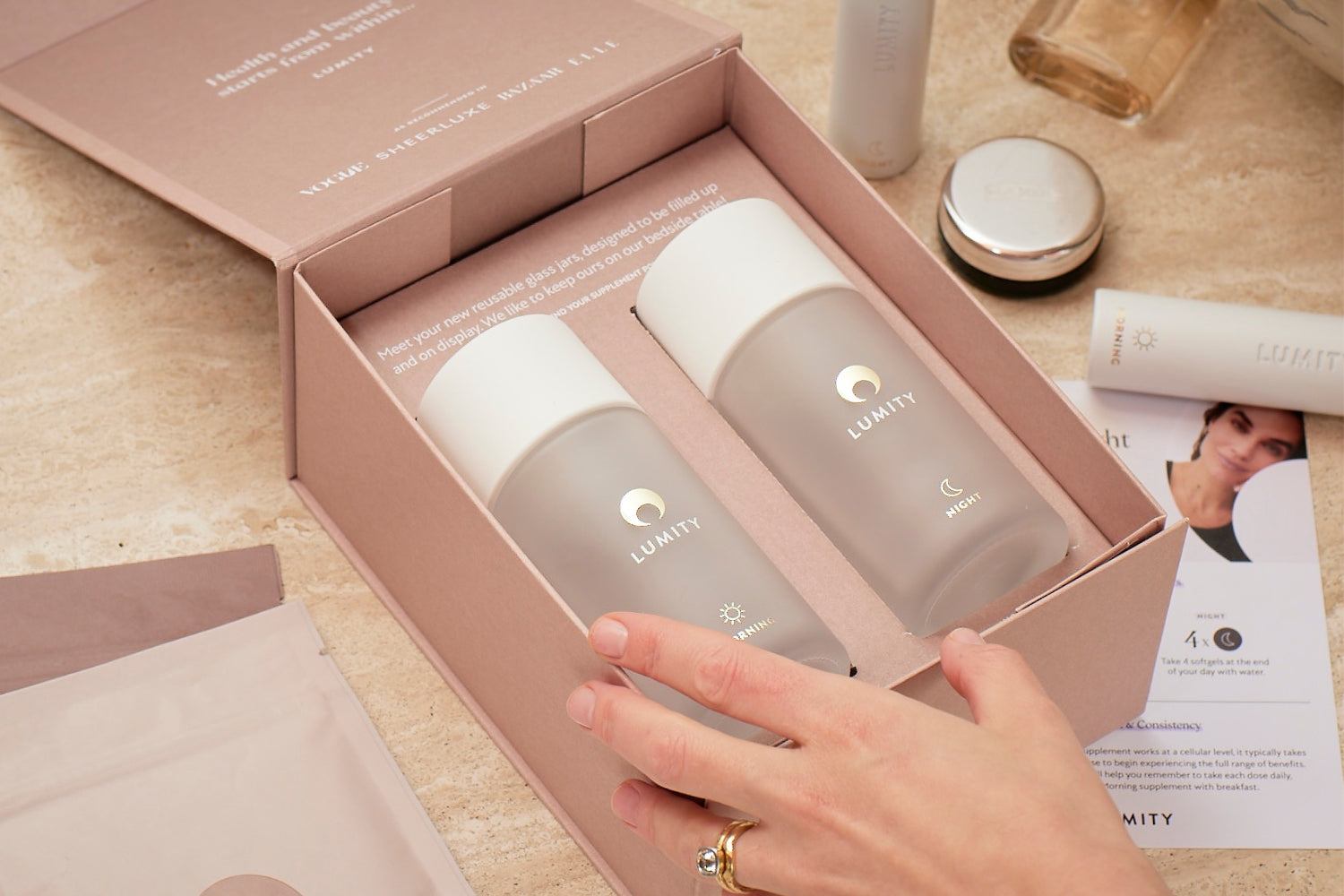 Open pink box holding two frosted Lumity bottles with white caps, surrounded by skincare and wellness items on a counter