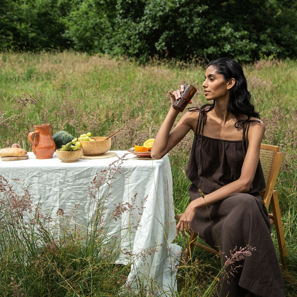 A woman in a brown sleeveless dress sits on a wooden chair in a grassy meadow, sipping from a brown glass. Beside her is a white-clothed table with a rustic spread of fruit, bread, and an orange pitcher, set against a lush, green backdrop of tall grass and trees.