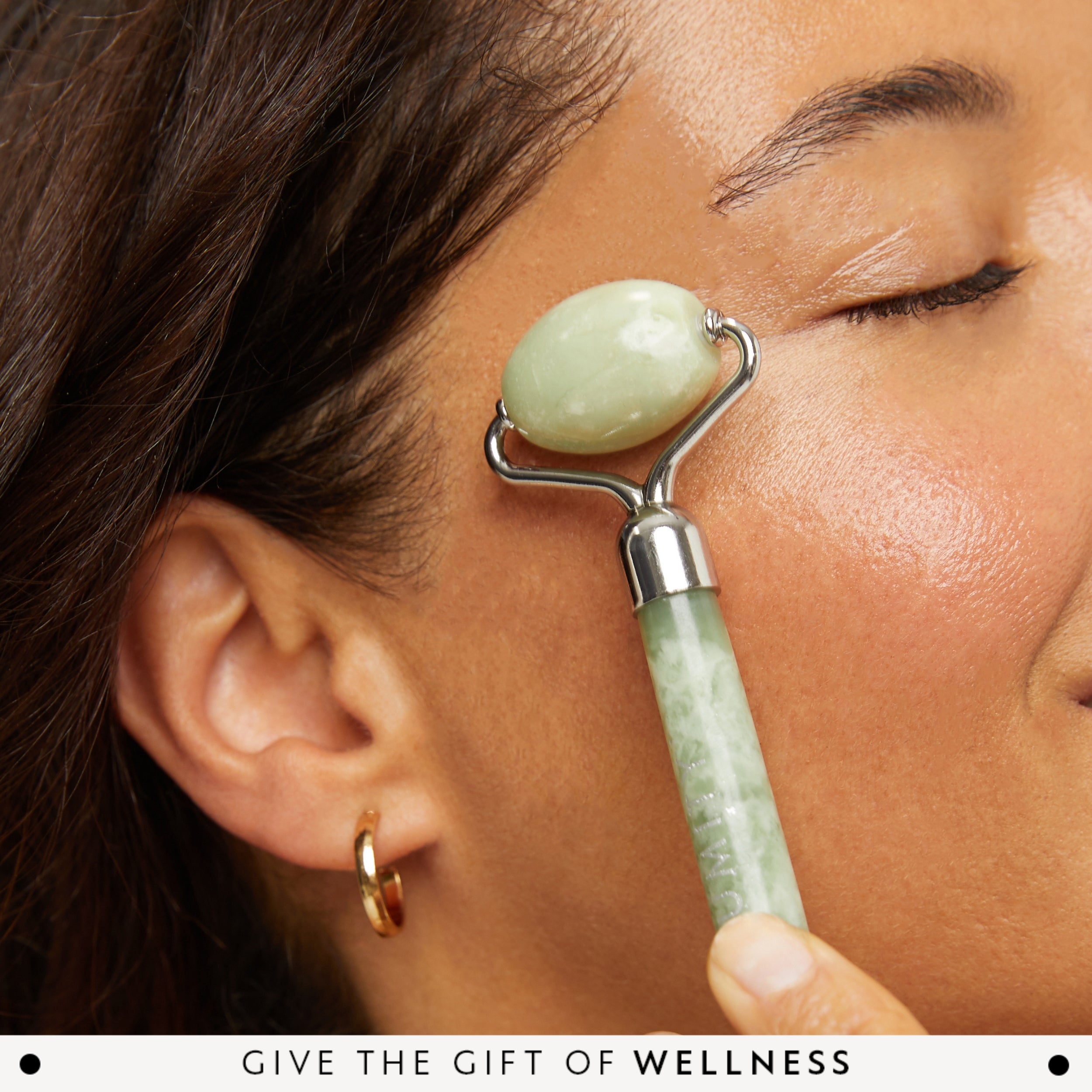 Person using a jade roller on their face with text 'Give the Gift of Wellness' at the bottom.
