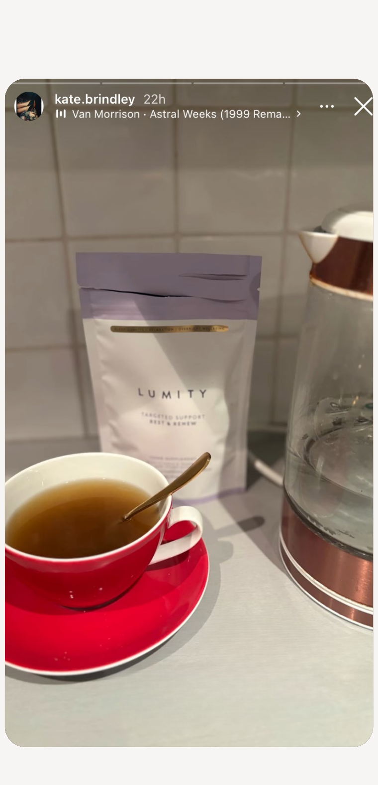 Red cup and saucer with tea and spoon beside Lumity supplement pouch and glass kettle on kitchen counter with tiled backsplash
