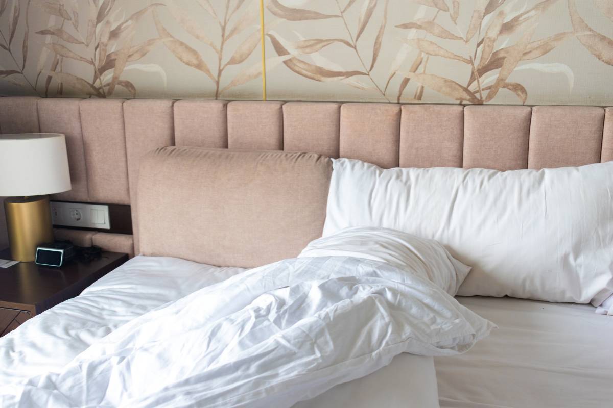 A bed with white bedding and two pillows in front of a tall, padded blush pink headboard. The wall behind the headboard features a beige botanical leaf patterned wallpaper. 