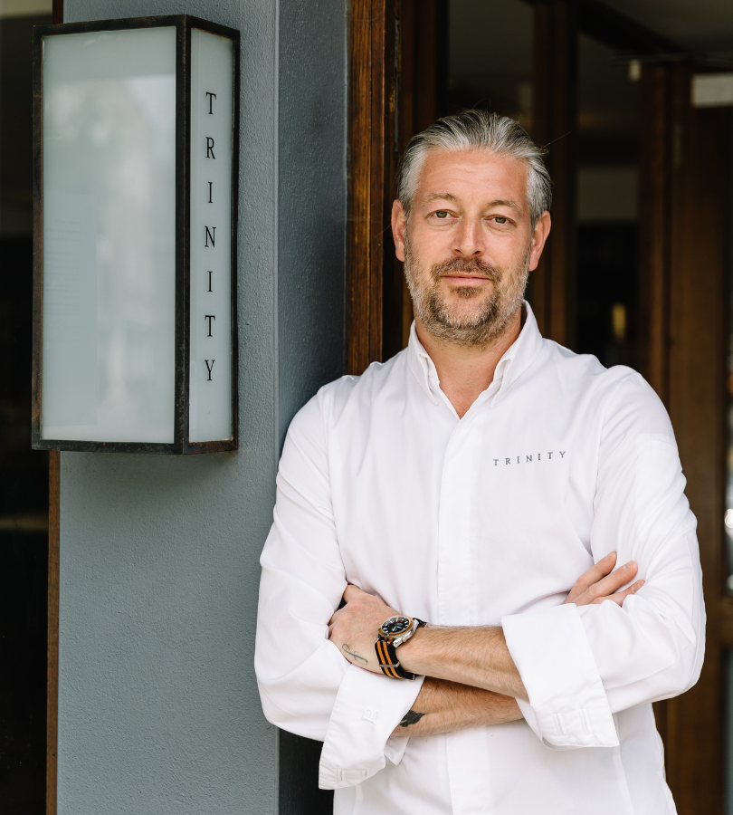 Man with salt-and-pepper hair and beard wearing a white chef shirt standing with arms crossed by a doorway
