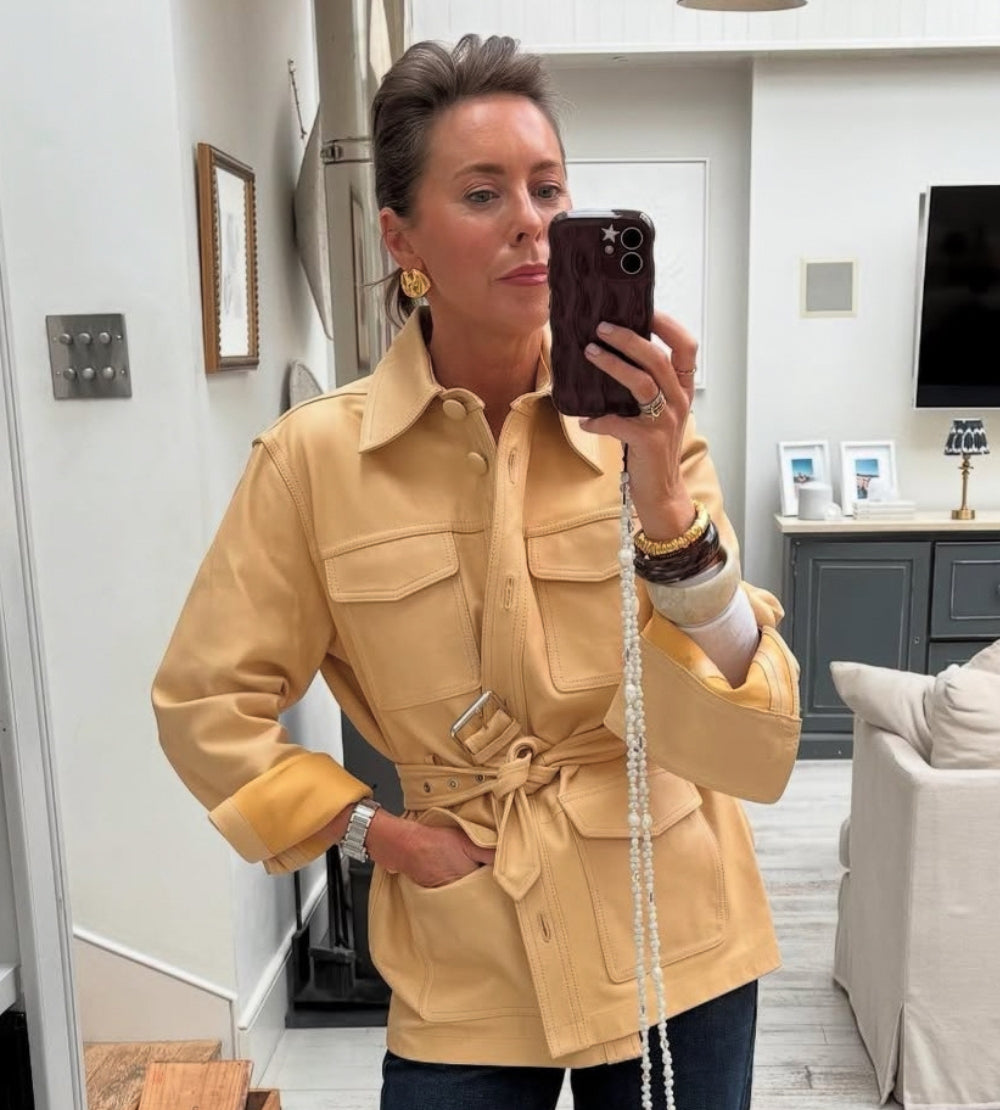 Woman taking a mirror selfie wearing a beige jacket in a living room.