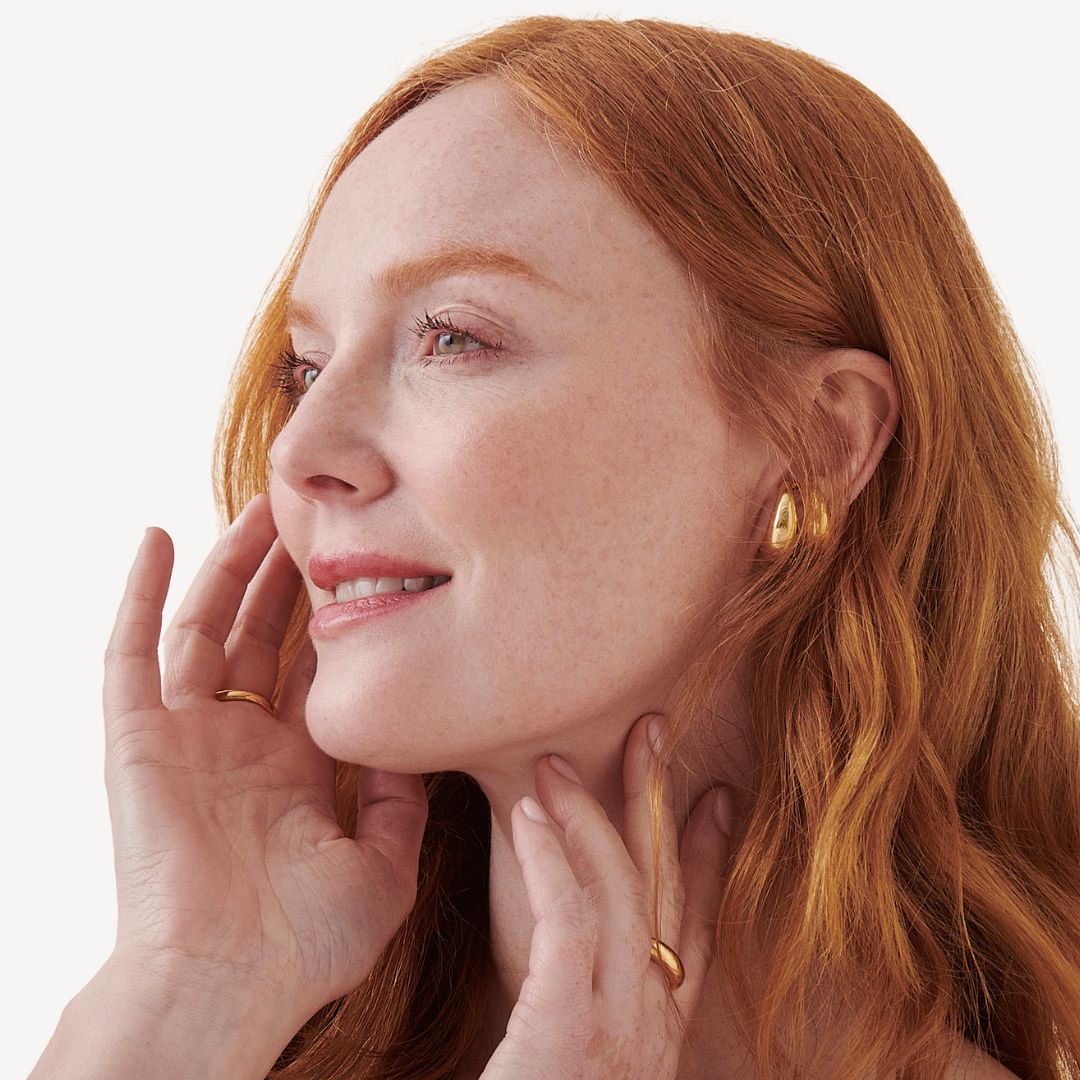 Red-haired woman with a gentle smile and closed eyes touching her face, wearing gold earrings and rings against a white background