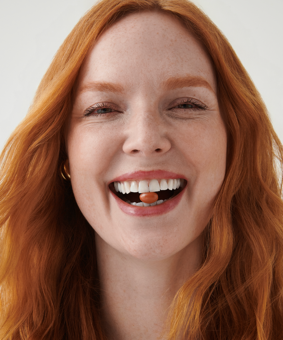 Close-up of a smiling woman with red hair holding an orange softgel between her teeth against a white background