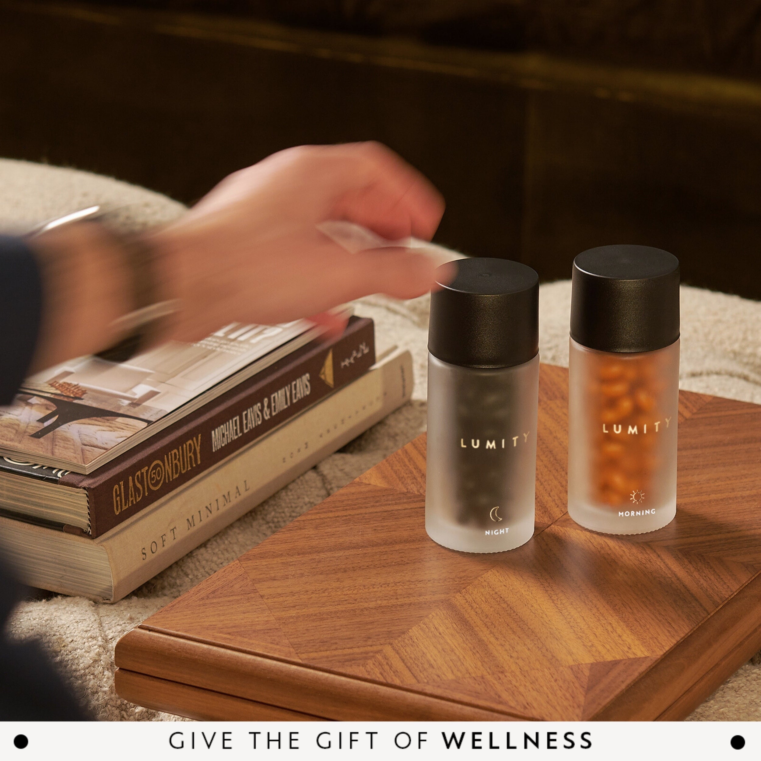 Two LUMITY bottles on a wooden table with books and hand reaching forward.