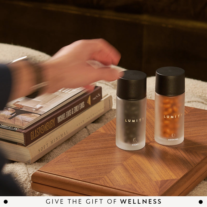 Two LUMITY bottles on a wooden table with books and hand reaching forward.