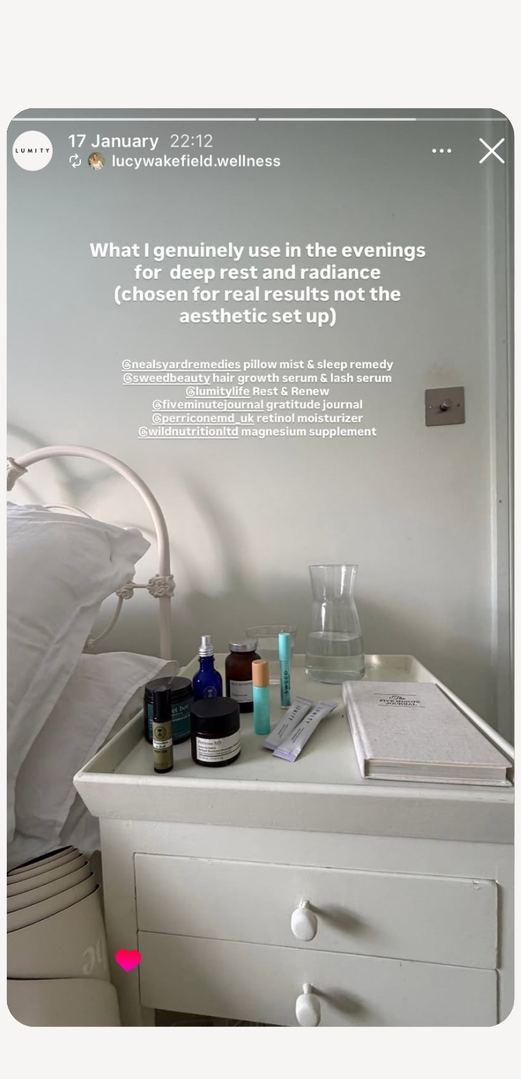 Nightstand with skincare and wellness items, glass carafe, journal, and white bed frame in softly lit bedroom corner
