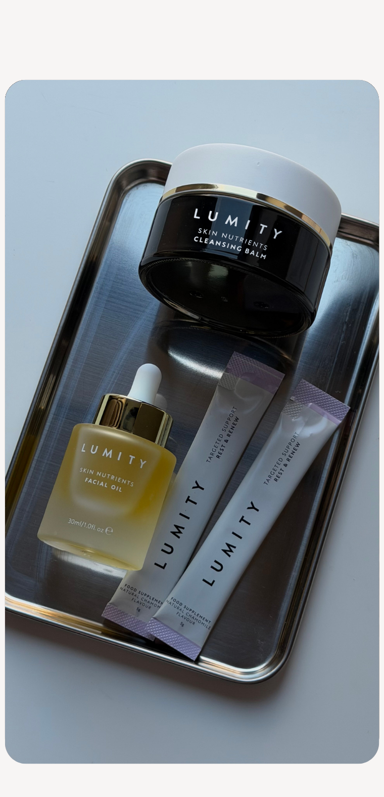 Lumity Skin Nutrients facial oil and cleansing balm with two Rest & Renew sachets on a stainless steel tray.