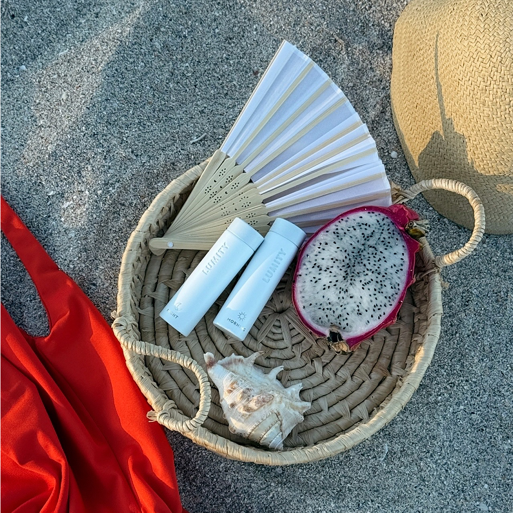 Basket with beach items including a fan, Lumity Travel Cases, dragon fruit, and a shell on a sandy surface.