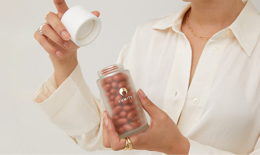 Woman wearing a white blouse holding an open glass jar of the Lumity Morning supplements